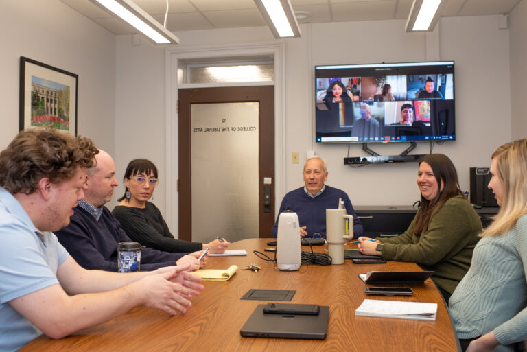 People sitting around a table having a meeting with a screen behind them showing people on a zoom call.